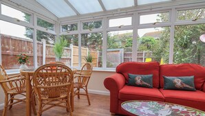 A conservatory with a red sofa, wicker chairs, a small table, and a view of a garden. - 31 Llys Ogwen (Prestatyn)