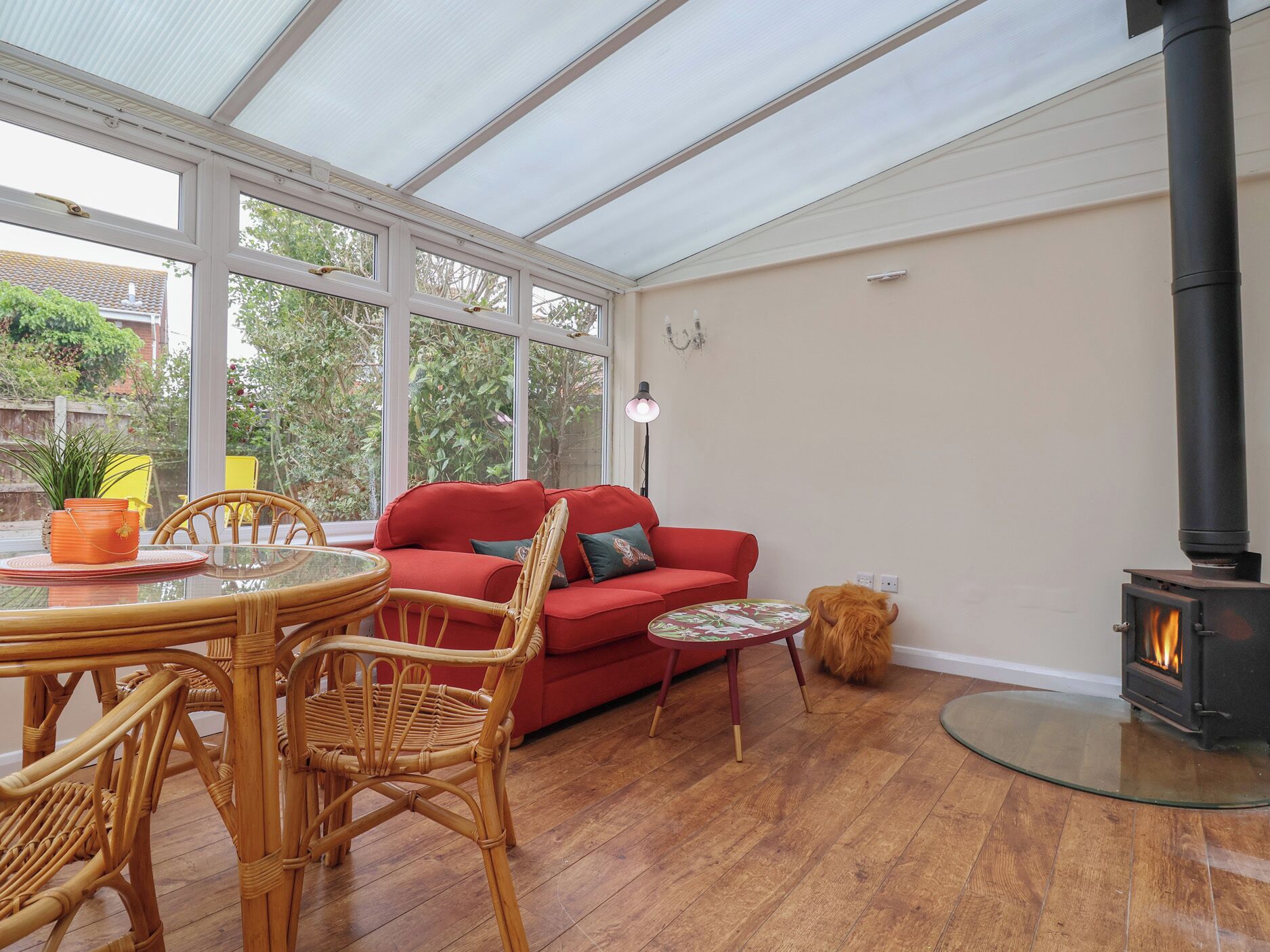 A cozy room with a red sofa, wicker chairs, a glass table, a small round table, a glass stove, and large windows.