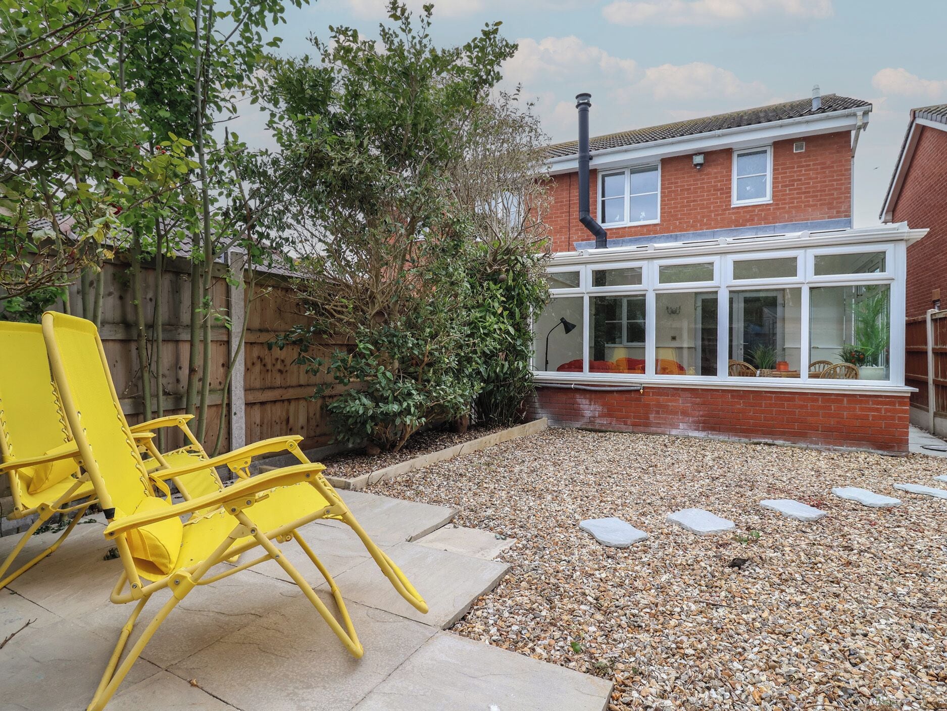 A patio with yellow chairs, a wooden fence, and a brick house with a window.