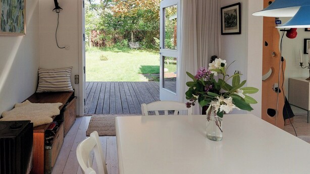 A room with a white table, a bench with a fur throw, a vase with flowers, and a view of the garden outside. - Cottage in woodland garden in natural harmony (Asnæs)
