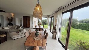 Amplia sala de estar con una mesa de comedor de madera, asientos cómodos, un ventanal con vistas a un paisaje verde y una lámpara colgante.