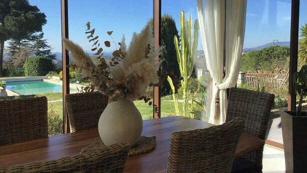 Un comedor con mobiliario de mimbre, un jarrón con plantas secas y vistas a una piscina y jardín.