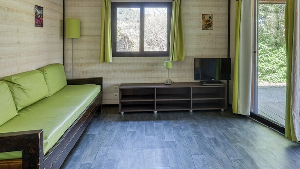 A room with a wooden ceiling, a green sofa, a TV on a stand, and a window with curtains. - Quiet Detached Chalet for Five (Sollies-Toucas)