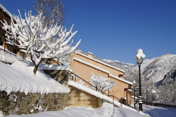 A snow-covered building with a stone wall, a staircase, and a street lamp. - Peaceful Alpine Triplex Escape (Guillaumes)