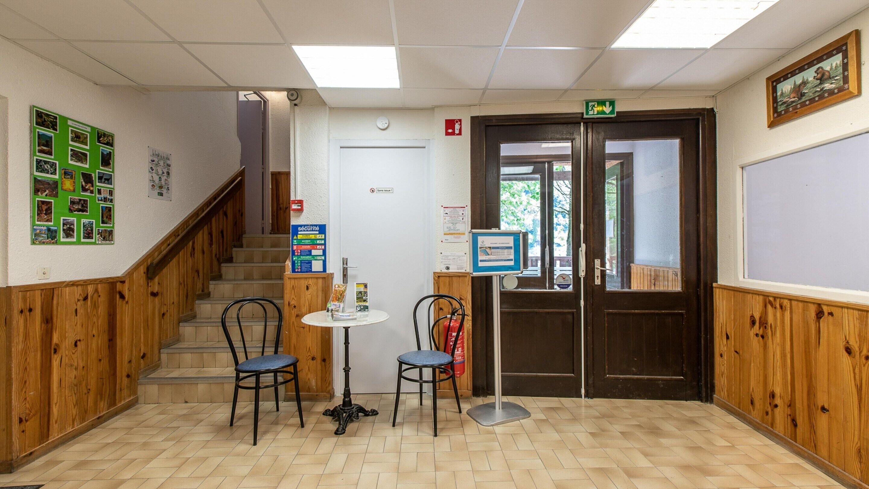 A reception area with a staircase, two chairs, a small table, a fire extinguisher, and a door with an emergency exit sign.
