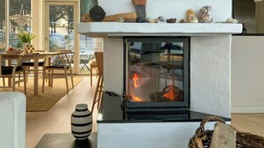 A modern living room with a fireplace, a dining area with a table and chairs, and a large window offering a view of the outdoors. - Restored fisherman's house on cliff in Thy (Hanstholm)