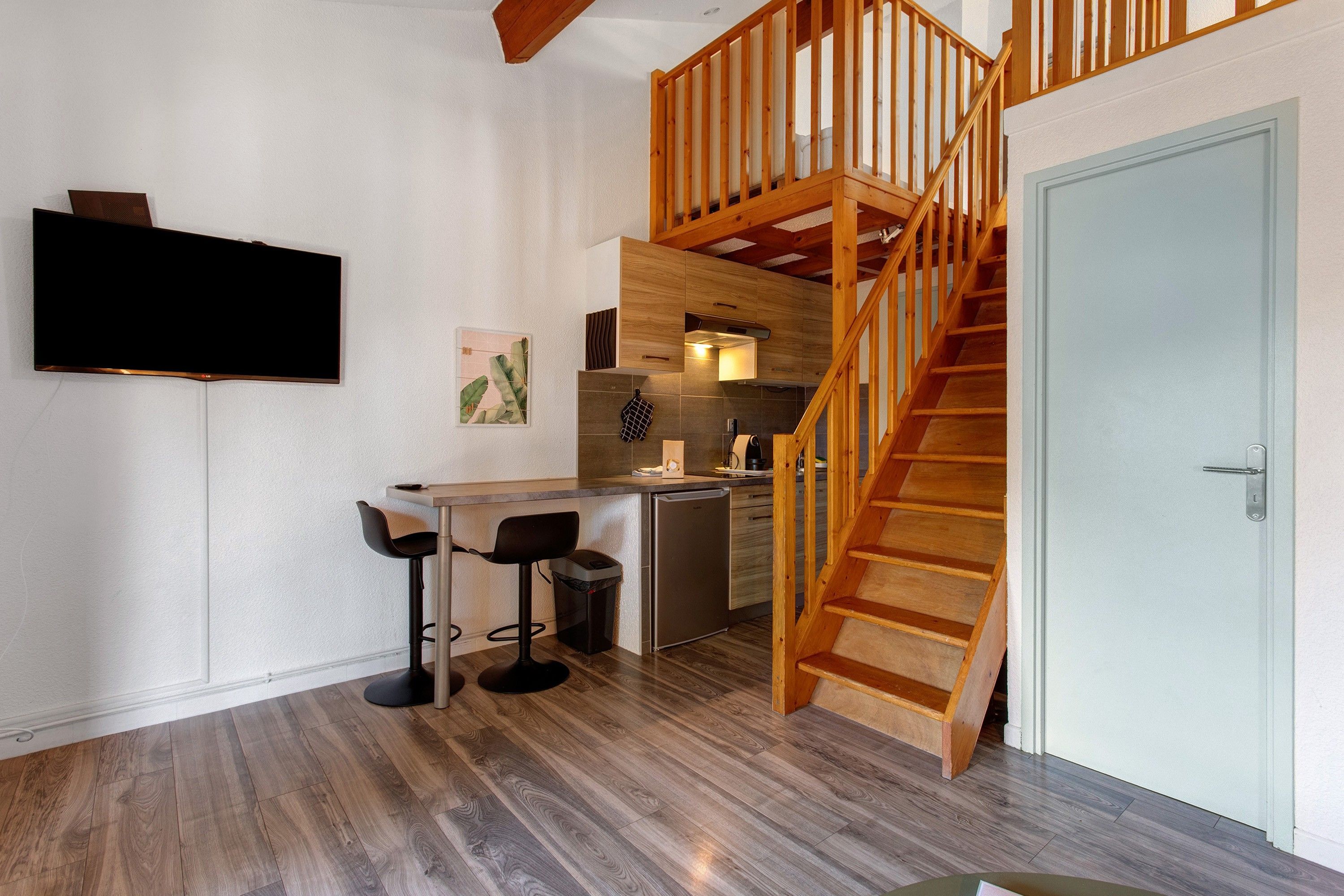 Een moderne keuken met een houten trap, een eethoek met barstoelen, en een flatscreen televisie aan de muur.