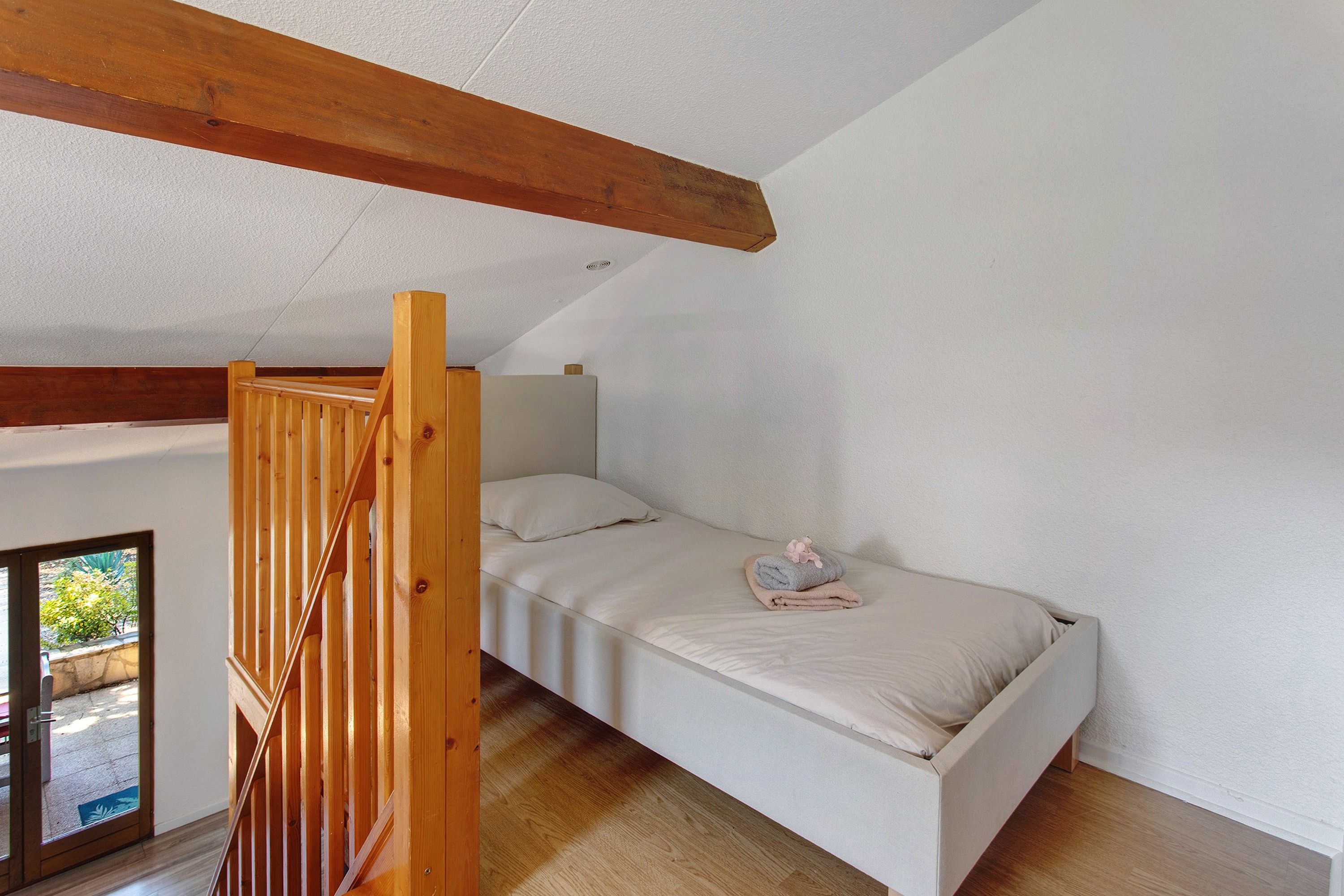 A bedroom with a sloped ceiling, a single bed, a wooden staircase, and a view through a door.