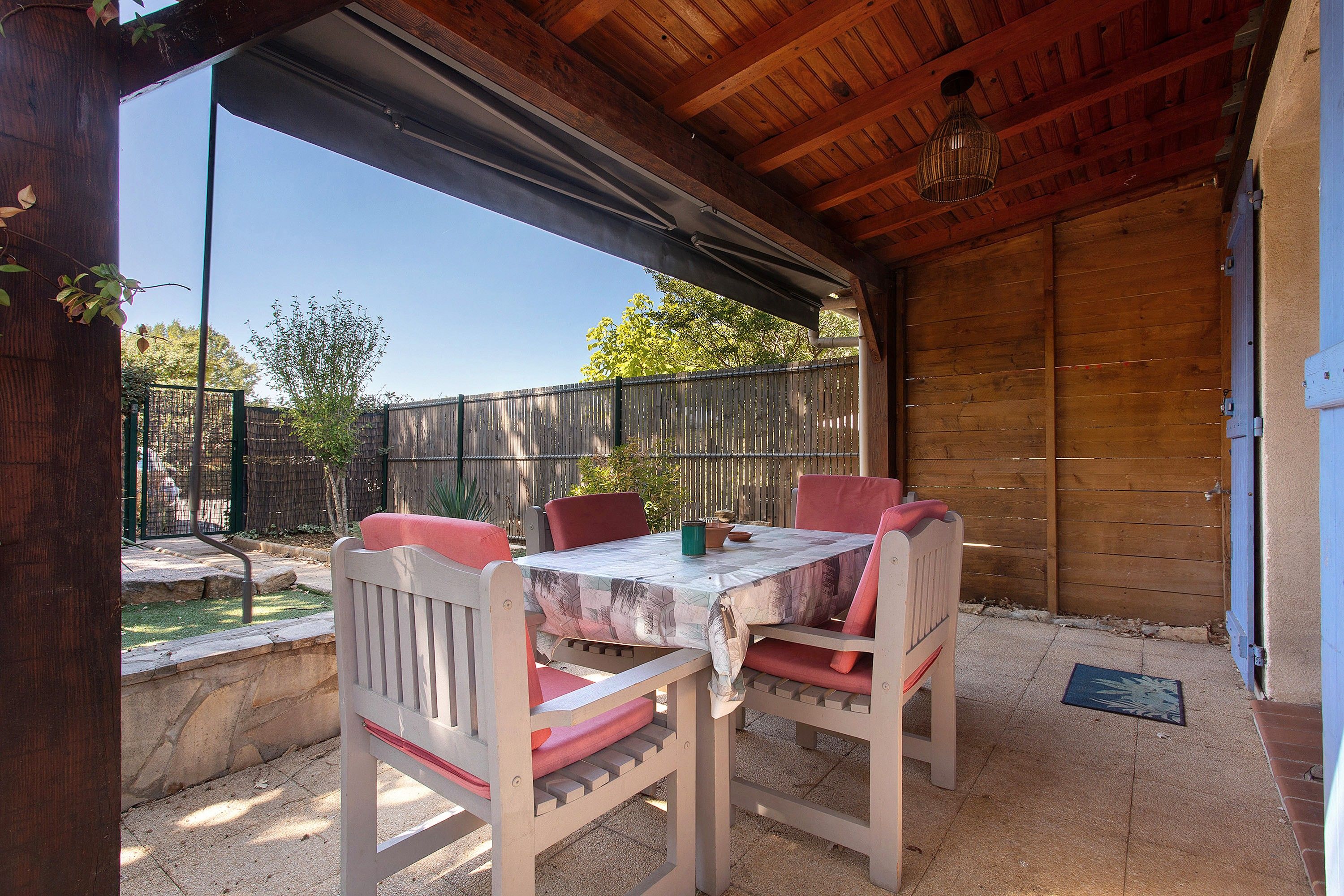 Un patio al aire libre con mesa y sillas, techo de madera y vista a un jardín cercado.