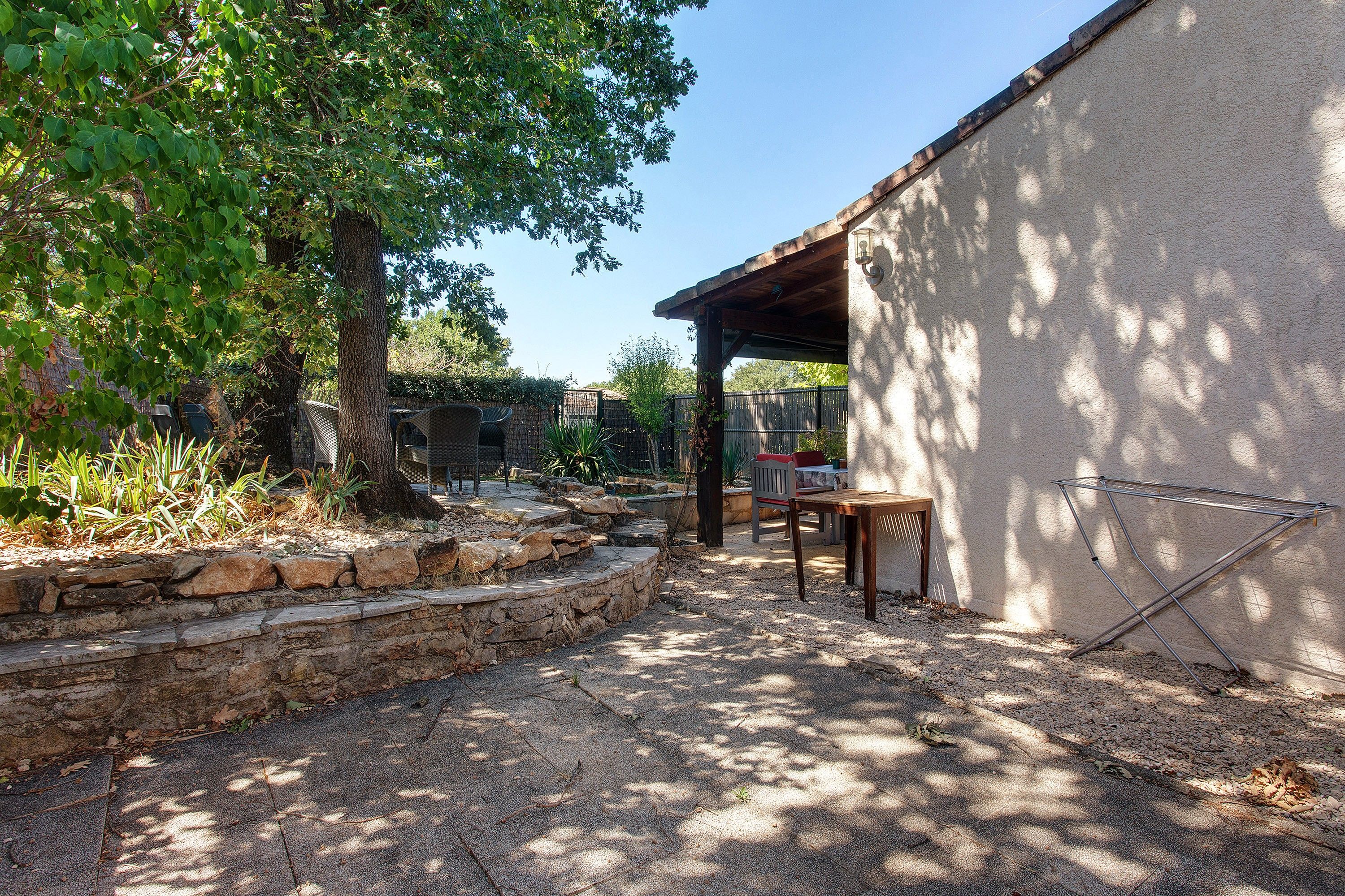 Un espace patio agrémenté d’un escalier en pierre, d’une table et de chaises, le tout entouré de verdure et d’arbres.