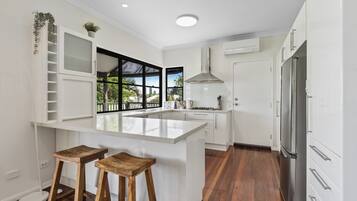 A modern kitchen with white cabinetry, a central island, two wooden stools, a stainless steel refrigerator, and a window with a view of palm trees.