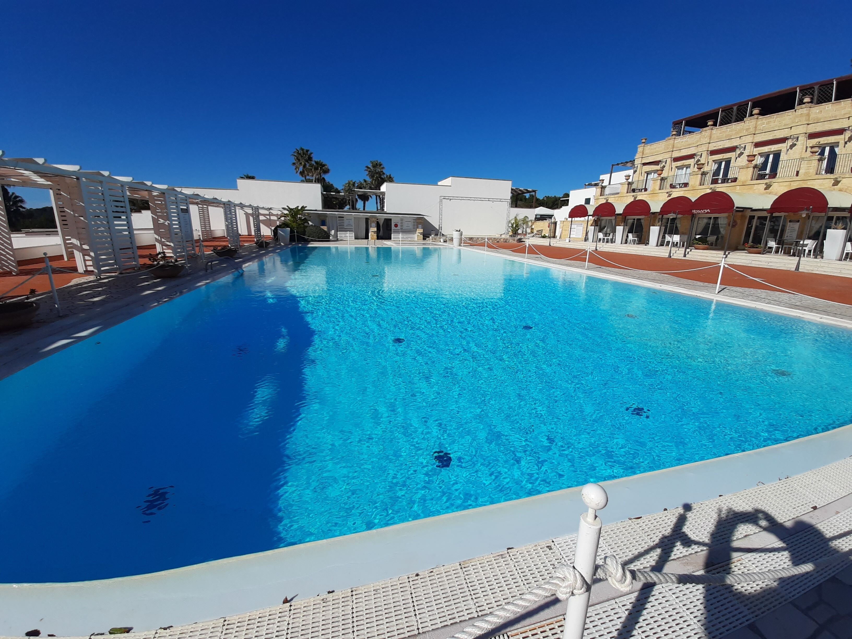 Piscina azul cristalina con un trampolín, rodeada de edificios y palmeras.