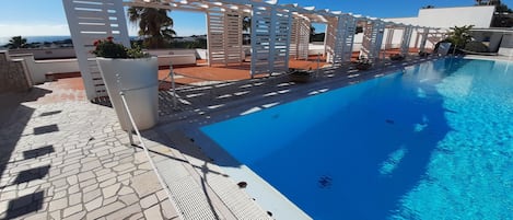 Una piscina con una pérgola de madera blanca, suelo de piedra y cielo azul despejado.