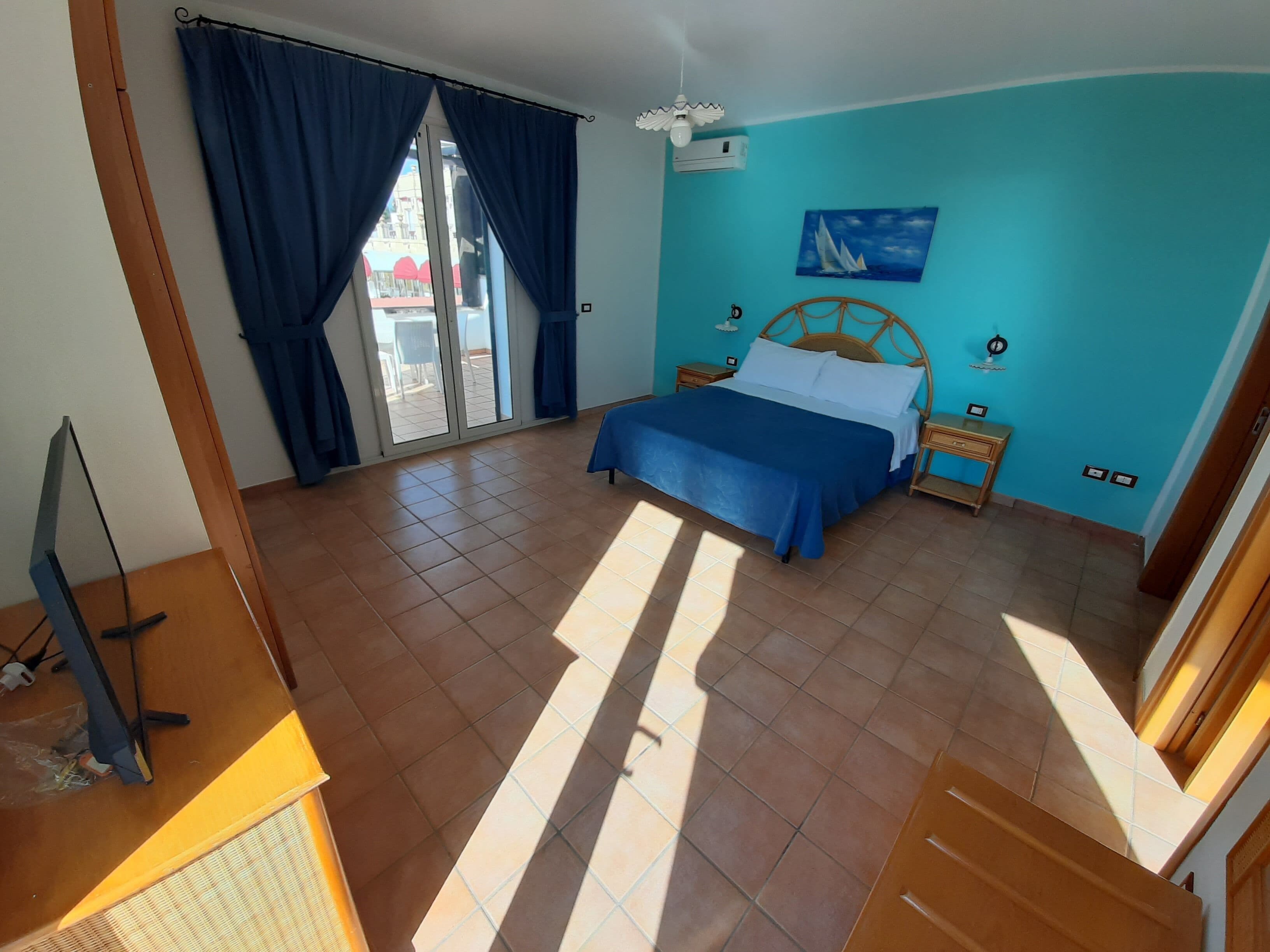 Une chambre à coucher comprenant un lit, un bureau, une chaise, une fenêtre avec des rideaux, un tableau au mur et une vue sur l’extérieur.