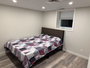 Una cama individual con una colcha estampada en una habitación con paredes blancas, una ventana con persianas y suelo de madera.