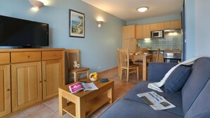 A living room with a TV, wooden cabinets, a sofa, a dining table, and a kitchen area with a microwave. - Holiday Home Near Lake Saint-cassien (Tourrettes)