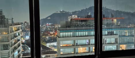 A room with a large window overlooking a cityscape at night, featuring modern buildings and a mountain in the background.