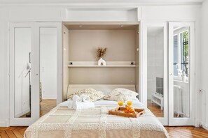 Quarto com cama, cabeceira de madeira, mesa de cabeceira com bandeja de croissants e suco de laranja, e vista para o exterior através de uma porta de vidro.