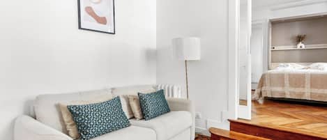 A white sofa with patterned cushions, a framed abstract artwork, a floor lamp, and a bed with a patterned bedspread.