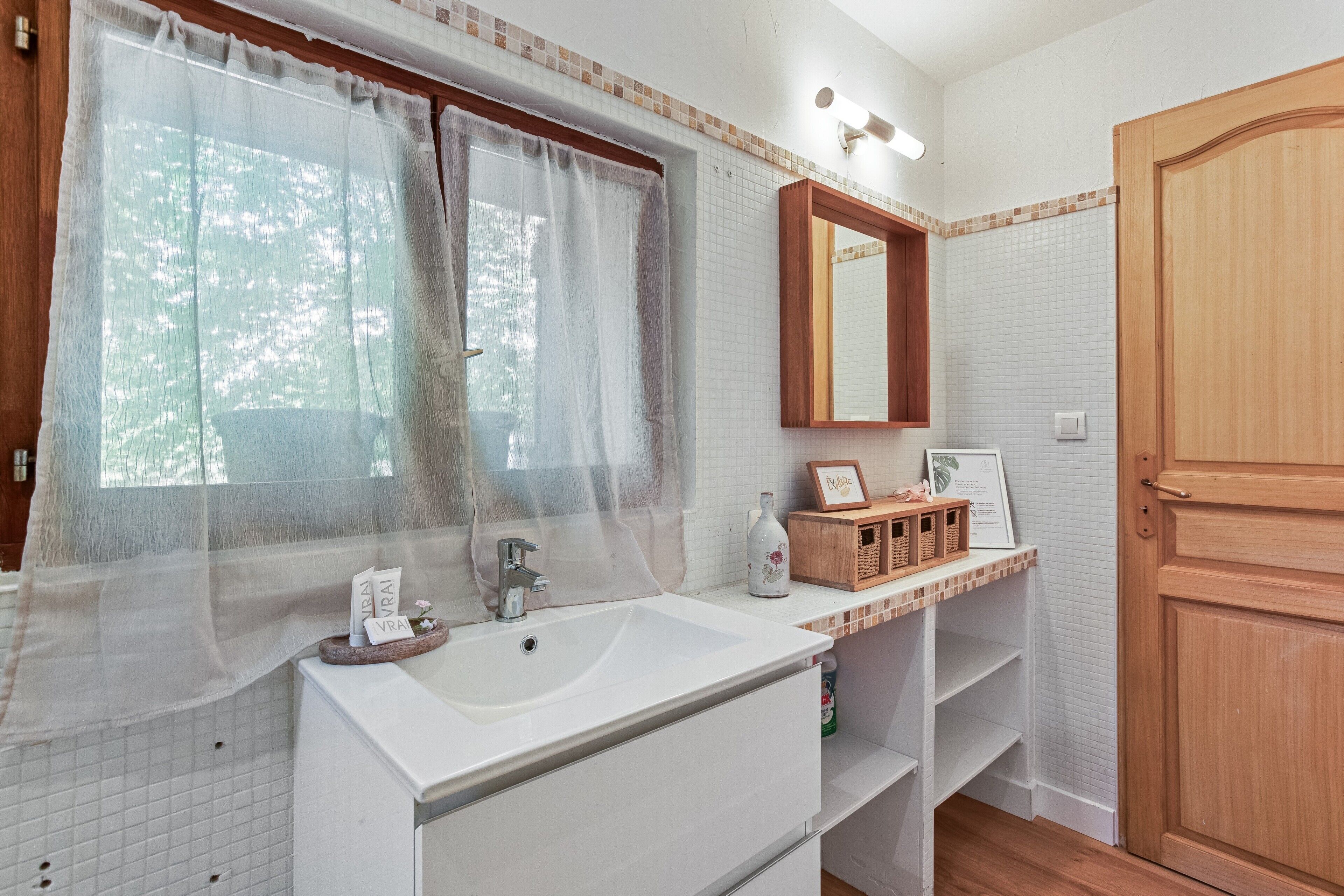 A bathroom with a large sink, a mirror, a window with sheer curtains, and a wooden door.