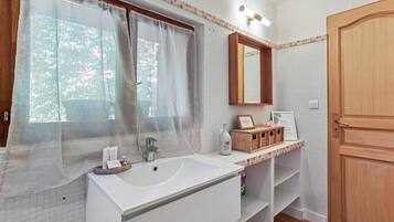 A bathroom with a large sink, a mirror, a window with sheer curtains, and a wooden door.