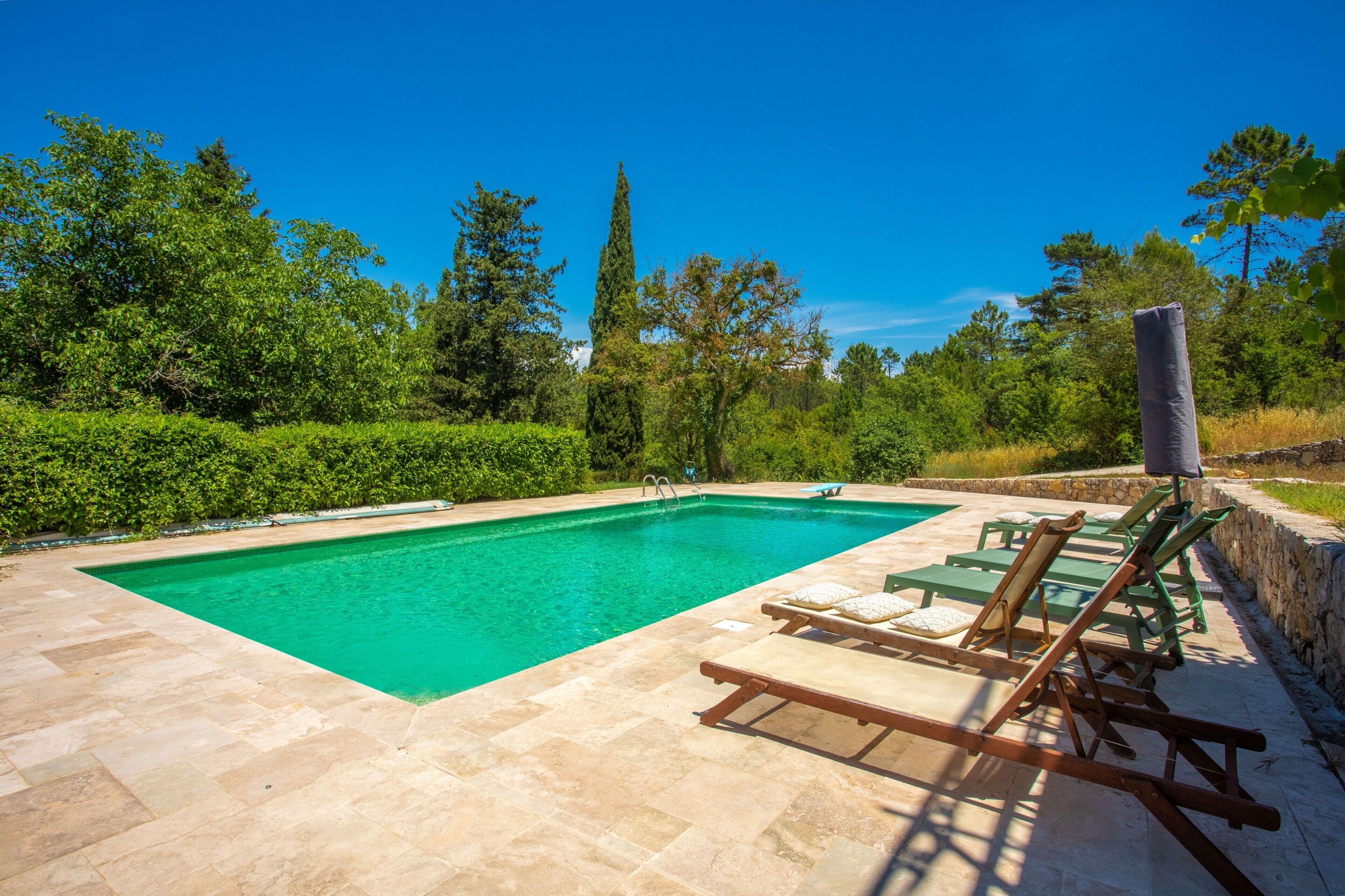 A rectangular swimming pool with clear water, surrounded by a stone deck. There are several green lounge chairs and a stone wall with a speaker.