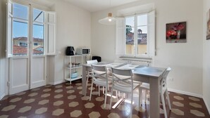 A dining area with a table and chairs, a white shelf with kitchen appliances, and a view of buildings through open windows. - Holiday Home Armonia Lucca (Lucca)