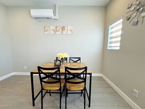 A dining area with a wooden table, four chairs, a vase with flowers, and a wall-mounted air conditioner.