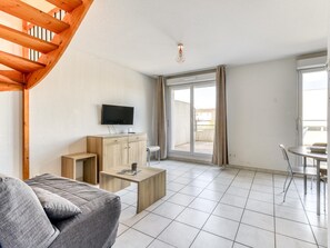 A modern living room with a sofa, a flat-screen TV, a wooden cabinet, a small table, and a balcony with a view. - Spacious Coastal Stay for Six With AC (Le Grau-Du-Roi)
