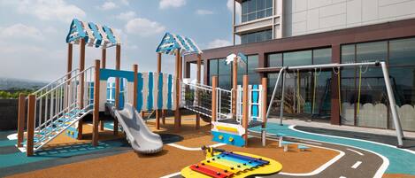 Zona de juegos infantiles con toboganes y estructuras de escalada, junto a un edificio con amplias ventanas.