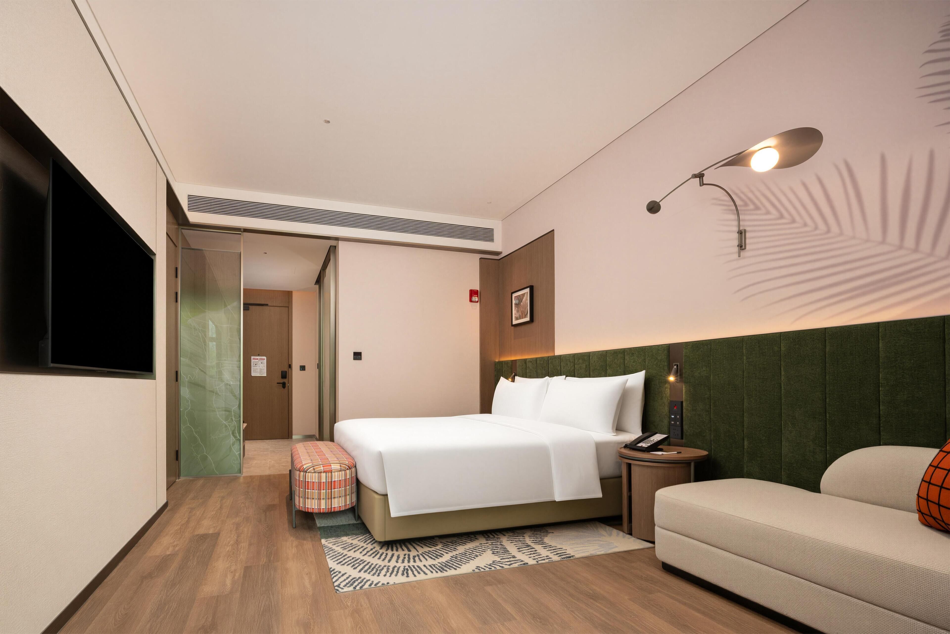 A hotel room with a bed, a bedside table, a lamp, a sofa, and a small stool.
