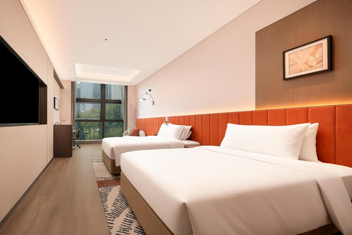 A hotel room with two beds, a large window, a flat-screen TV, and a framed picture on the wall.