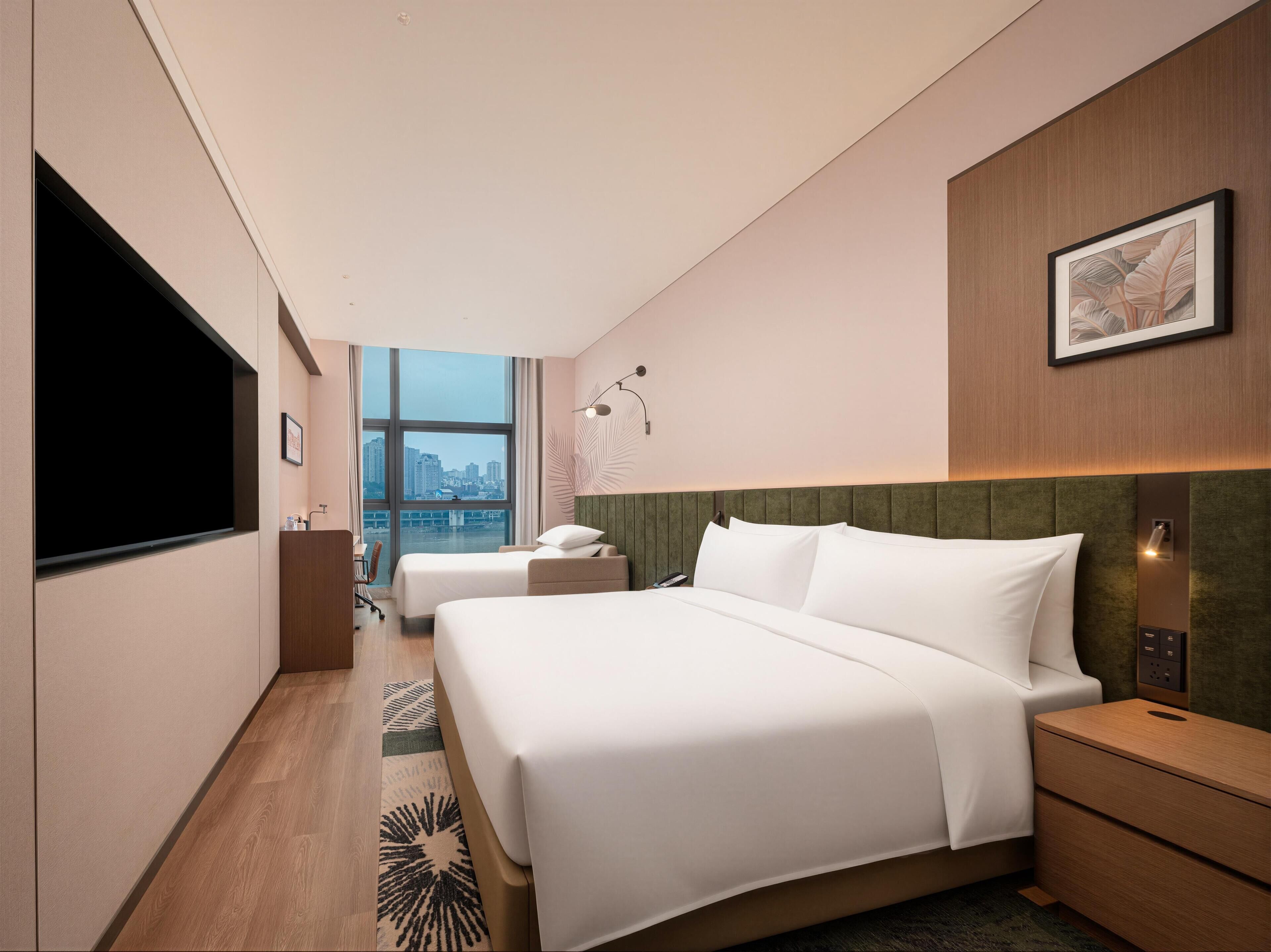 A hotel room with a large bed, a desk, a chair, a television, and a city view.