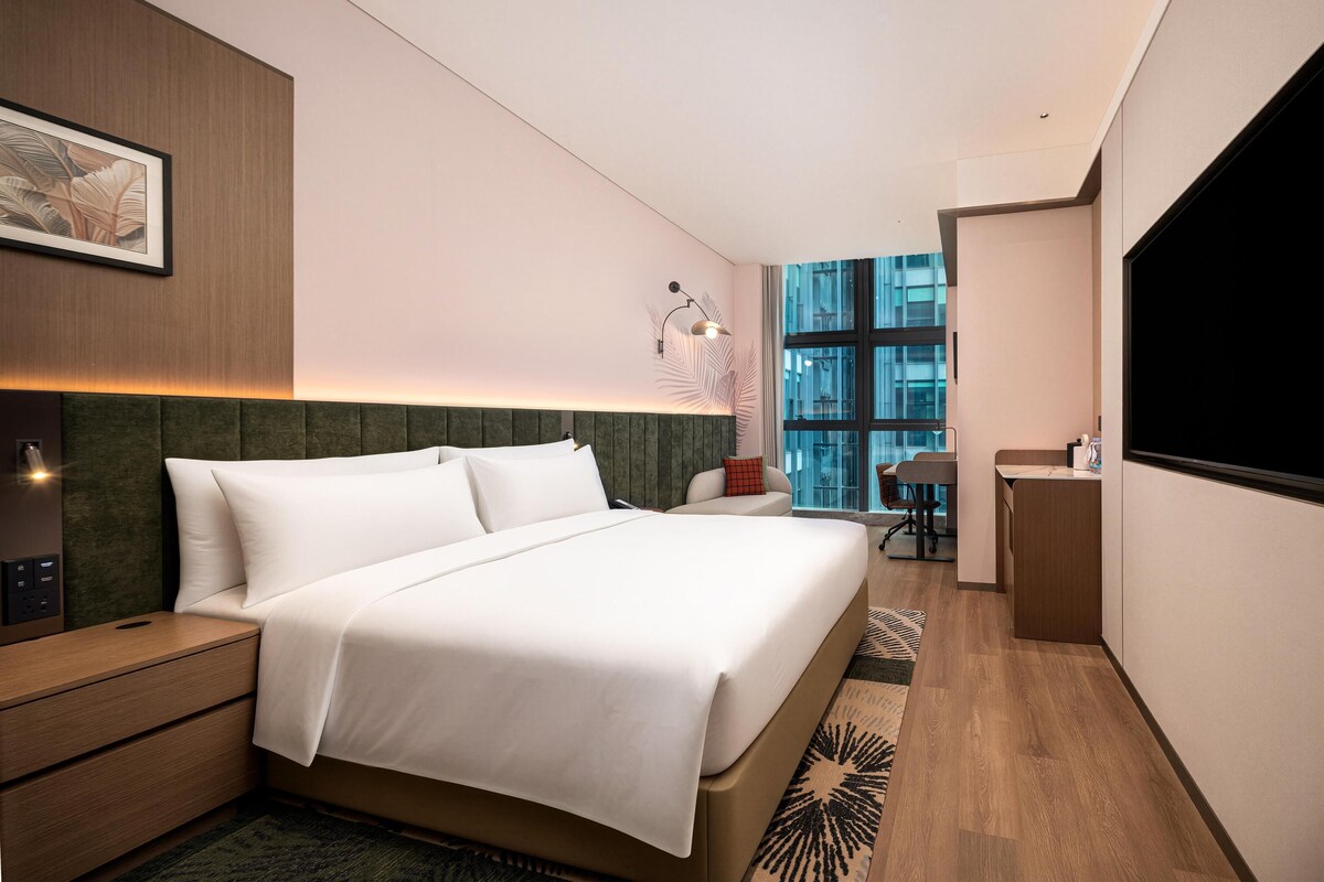 A modern hotel room with a large bed, bedside tables, a desk, and a large window offering a city view.