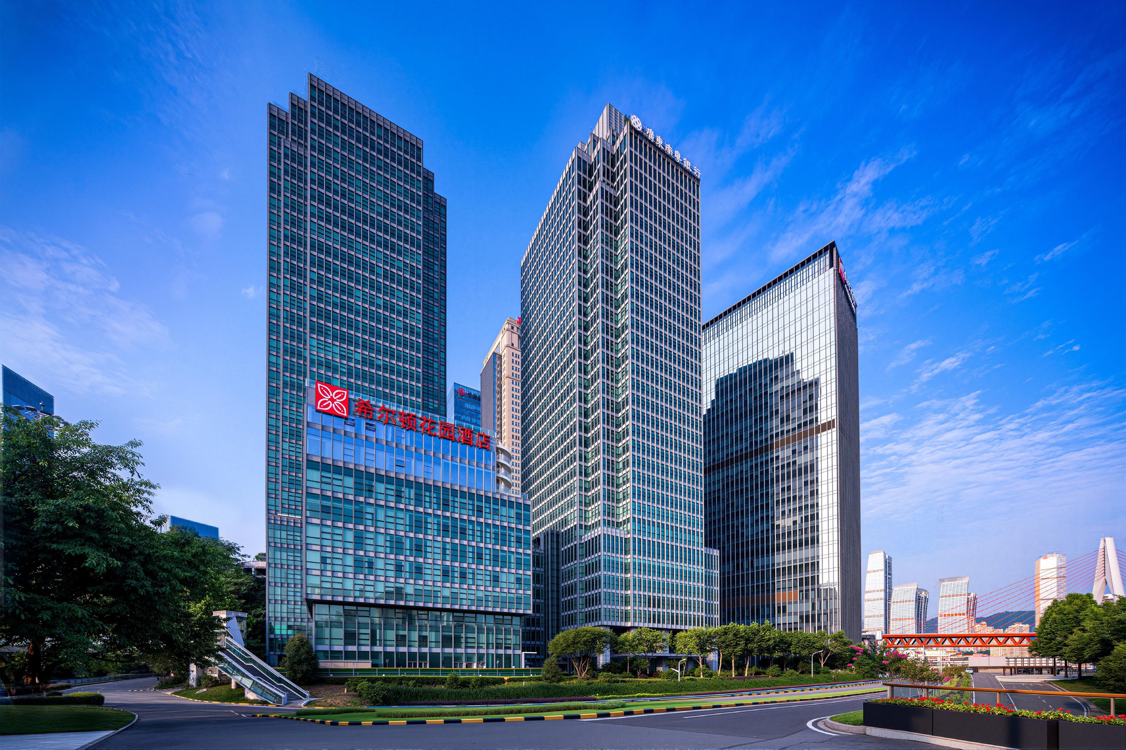 A modern hotel with a prominent red logo, surrounded by high-rise buildings and a clear blue sky.