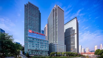 A modern hotel with a prominent red logo, surrounded by high-rise buildings and a clear blue sky.