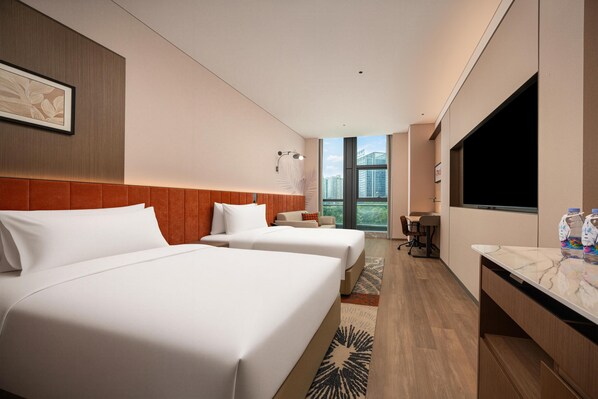 A hotel room with two beds, a TV, a desk, and a view of the city.