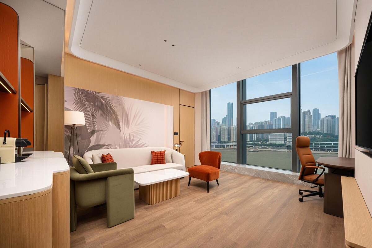 A modern hotel room with a city view, a sofa, a chair, a desk, and a large window.