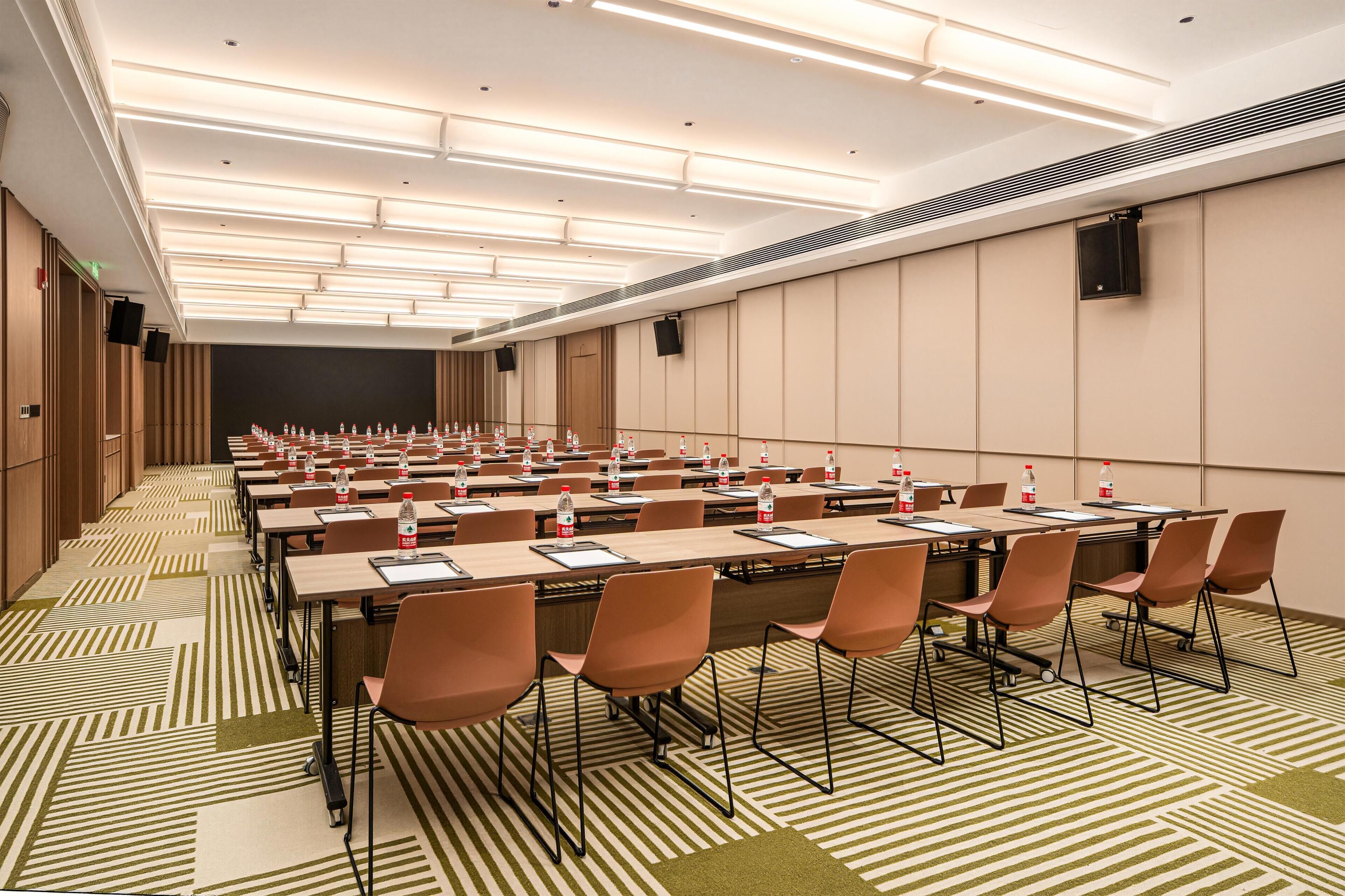 Un salón de conferencias con varias mesas y sillas, equipado con botellas de agua, y una pantalla grande al frente.