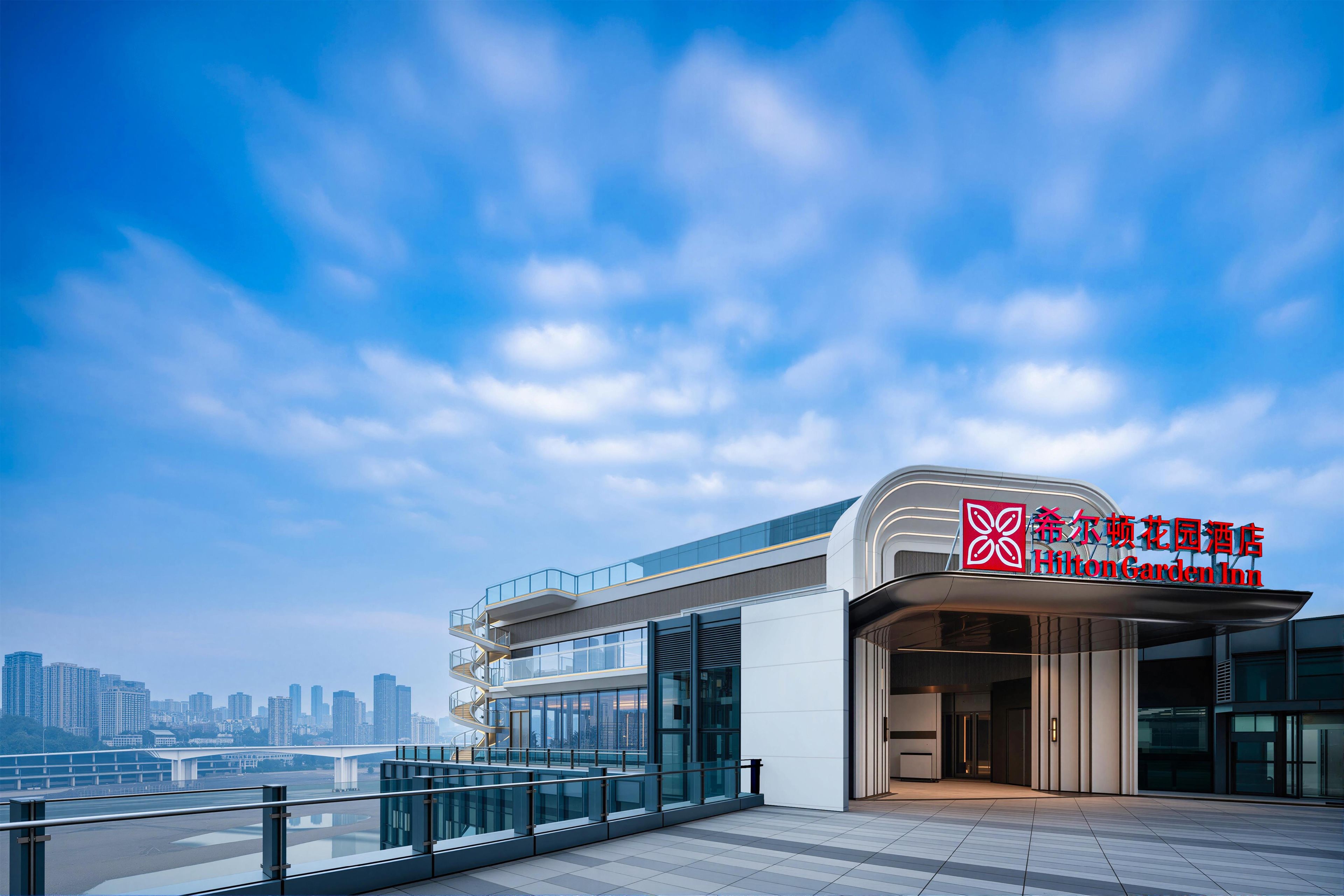 A modern hotel with a glass facade, a prominent sign for Hilton Garden Inn, and a view of the city skyline in the background.
