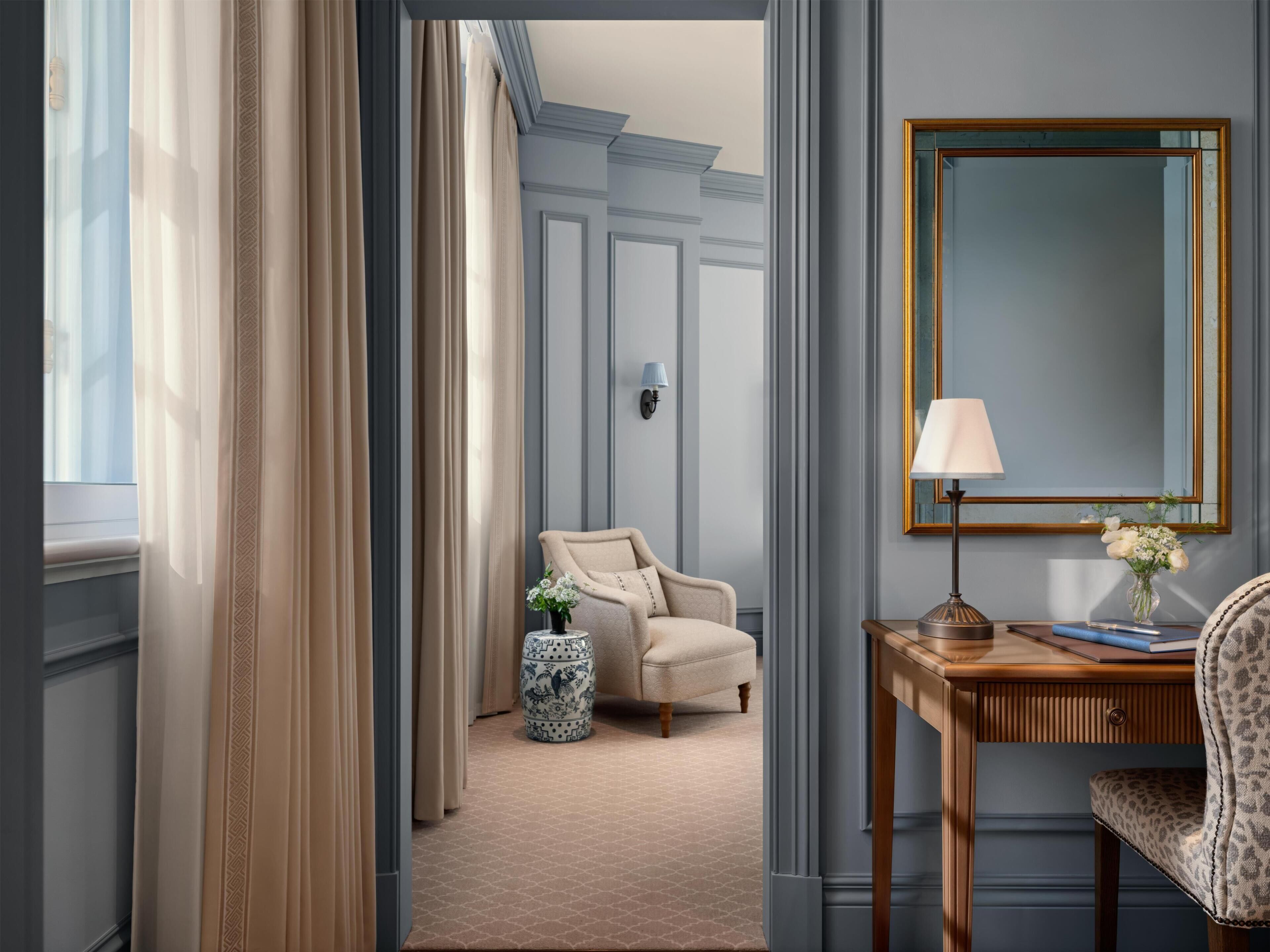 Habitación de hotel con un ventanal grande, una silla, una mesita con una lámpara y un espejo en la pared.