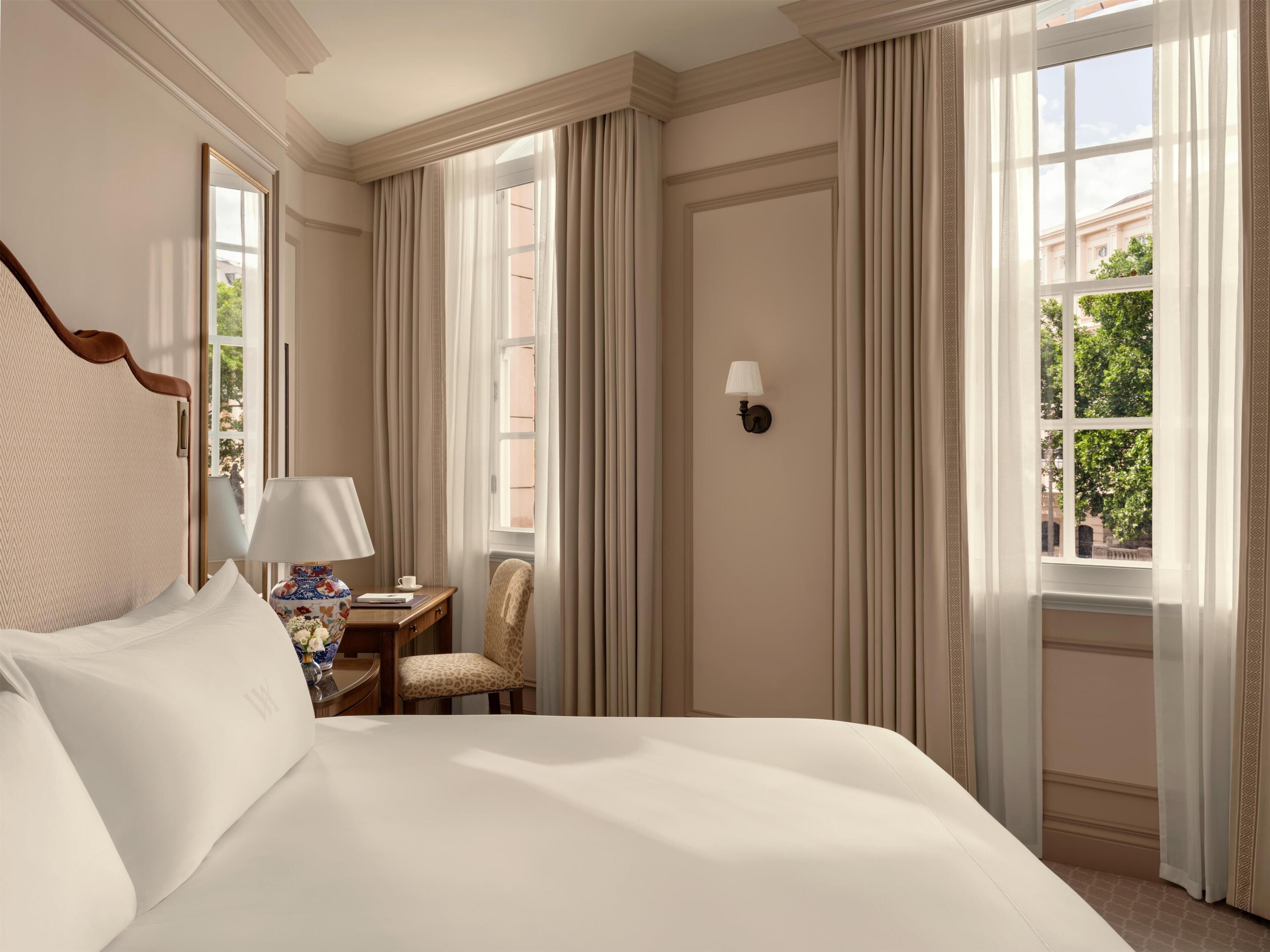 Habitación de hotel con una cama grande, un escritorio con lámpara, una silla y una ventana con cortinas.