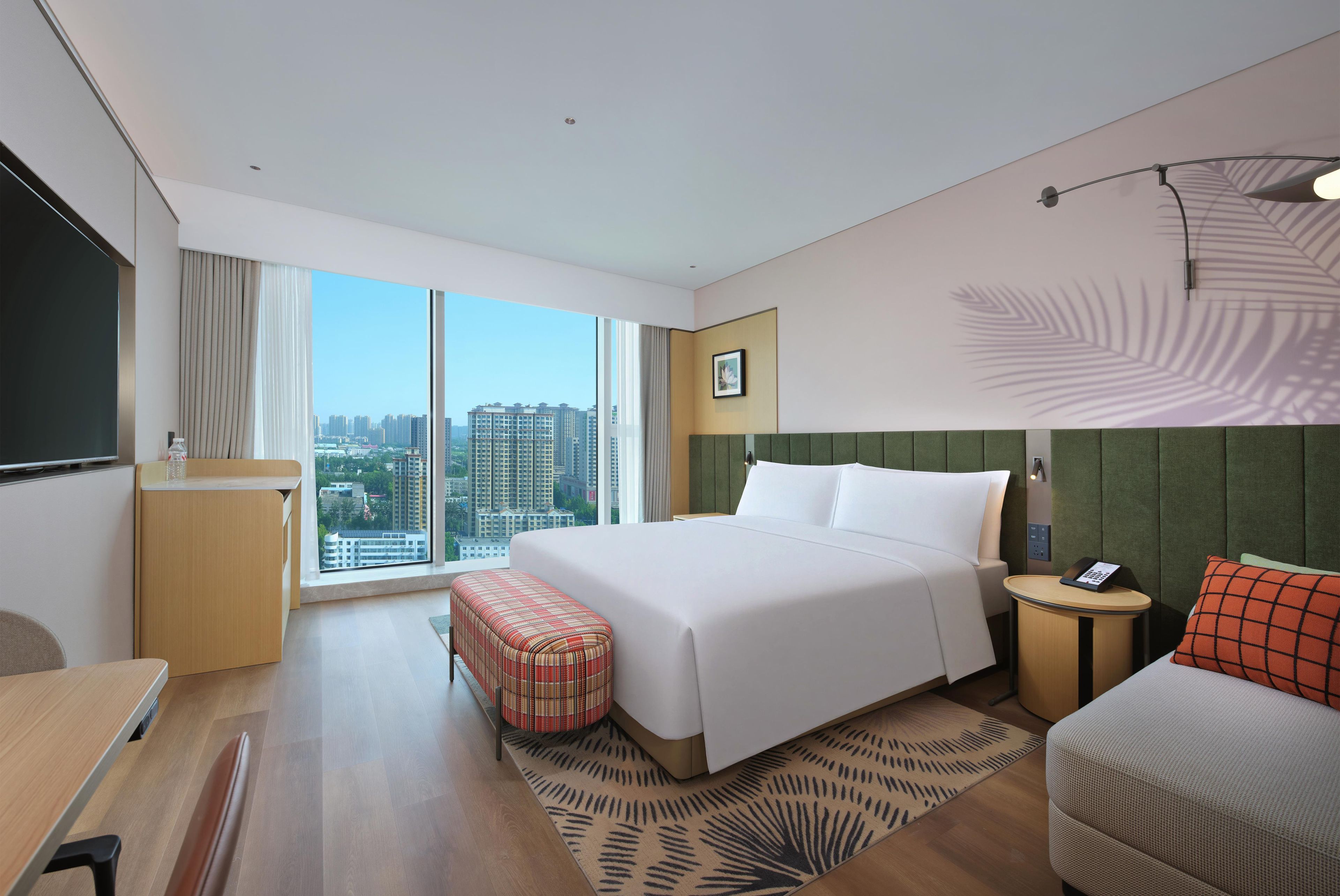 A hotel room with a large bed, a desk, a chair, a small table, a sofa, a large window with a city view, and a wall-mounted telephone.