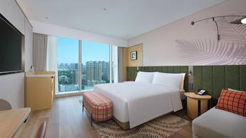 A hotel room with a large bed, a desk, a chair, a small table, a sofa, a large window with a city view, and a wall-mounted telephone.