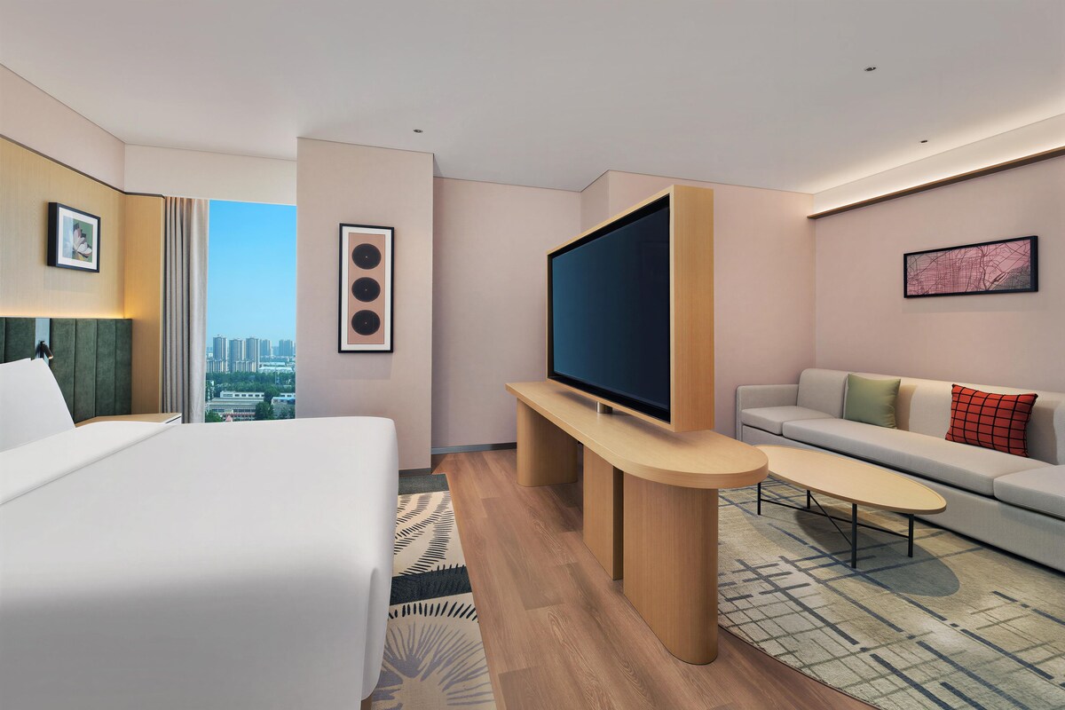 A hotel room with a bed, a TV on a stand, a sofa, and a city view.