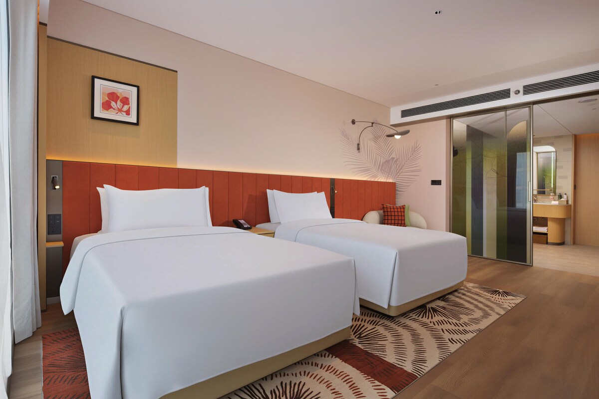 A hotel room with two beds, a wall-mounted artwork, a control panel, and a sliding door leading to a bathroom.