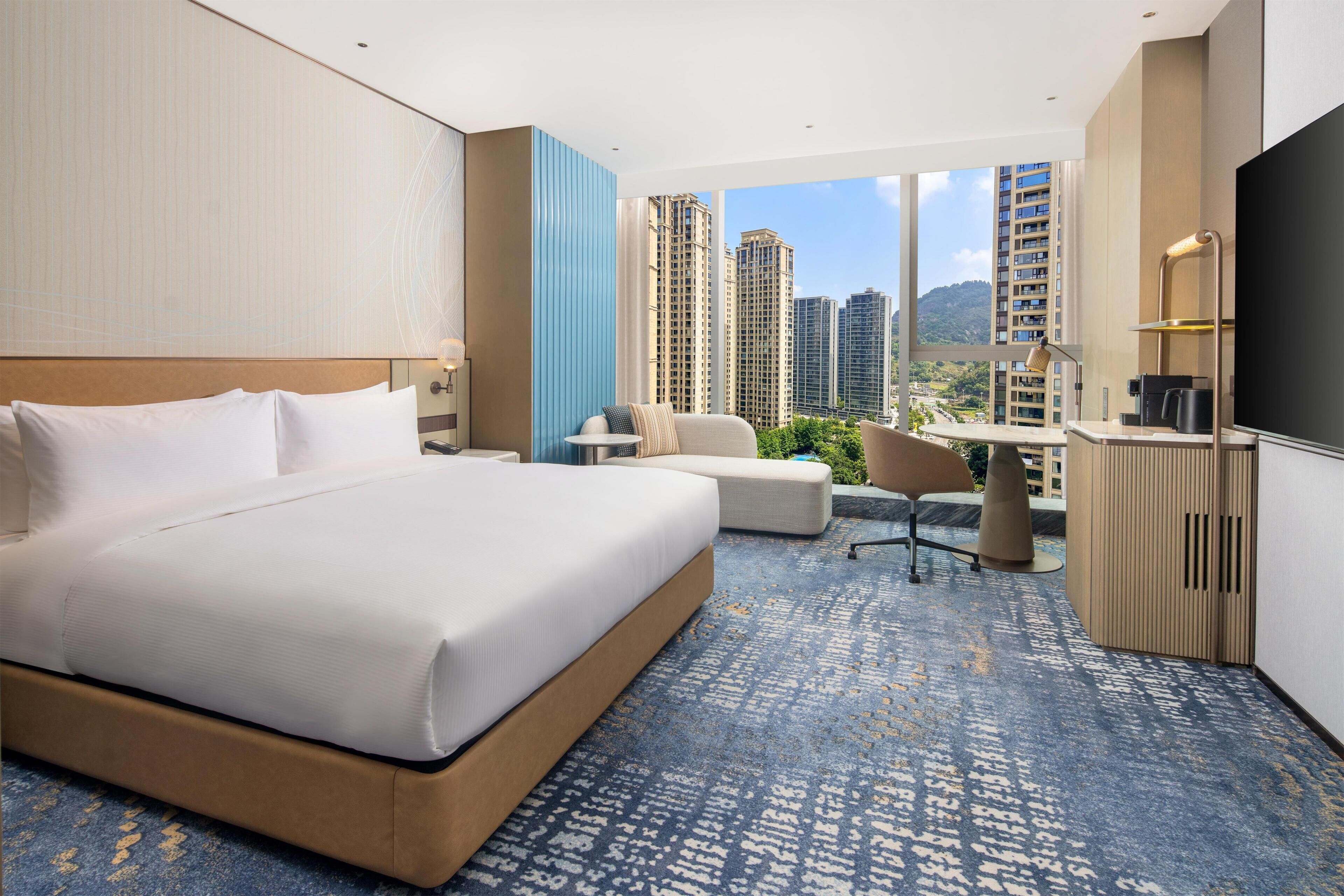 Habitación de hotel con una cama grande, un escritorio, una silla y vista a edificios altos.