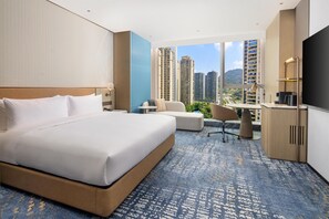 A hotel room with a large bed, a desk, a chair, and a view of tall buildings. - Hilton Wenzhou (Wenzhou)