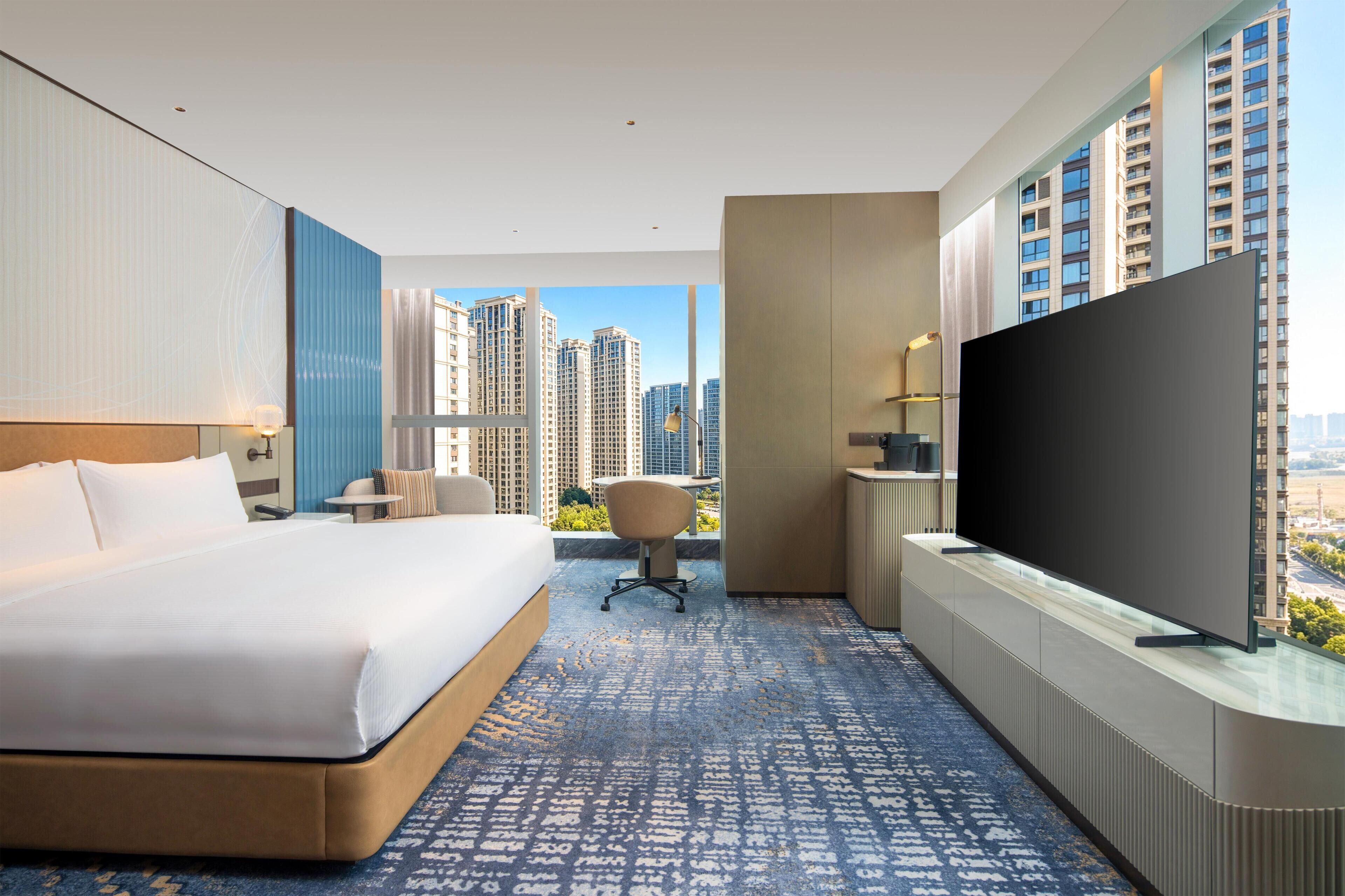 Habitación de hotel con una cama grande, un escritorio, una silla, un televisor y vista a la ciudad.