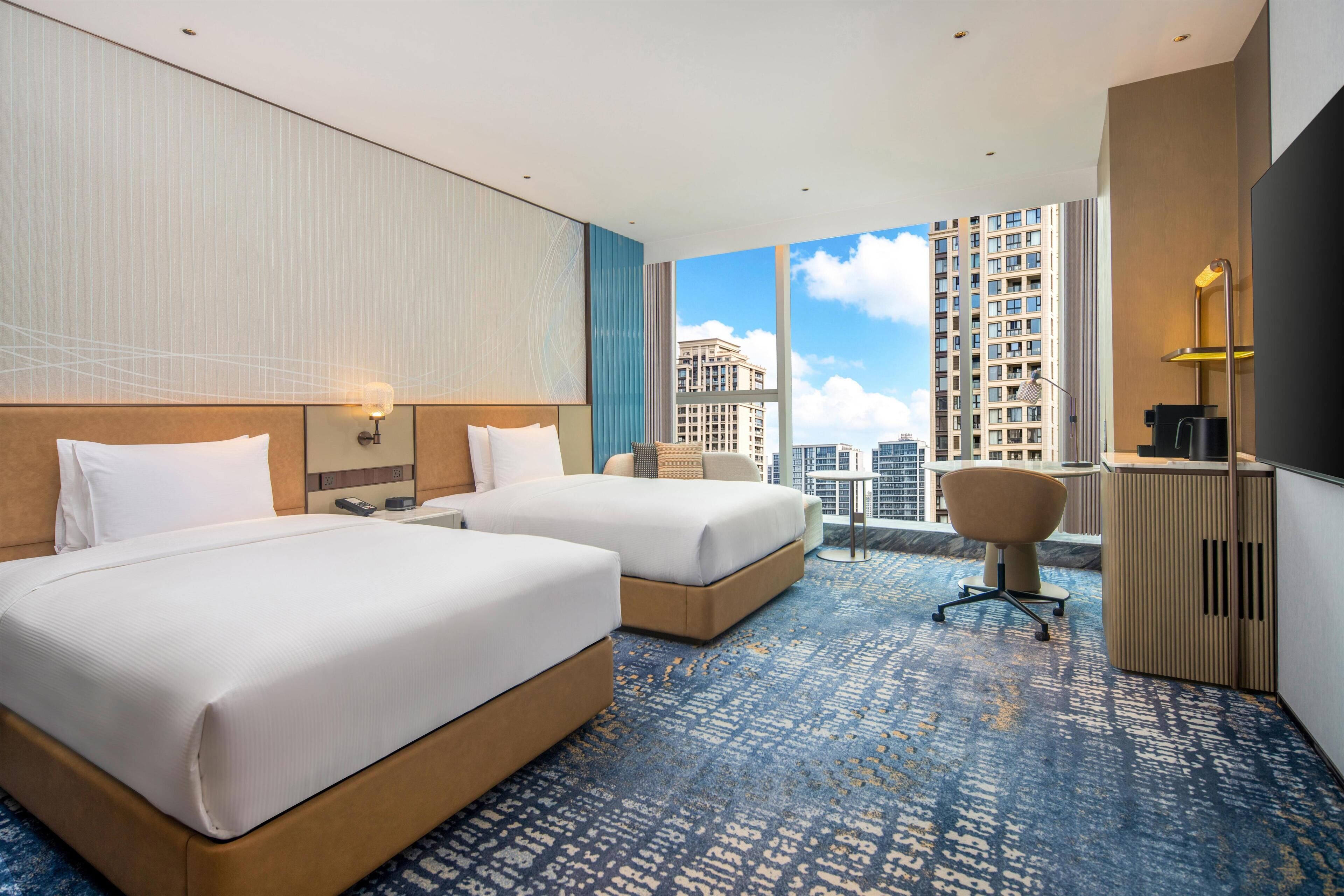 A hotel room with two beds, a desk, a chair, and a view of tall buildings.
